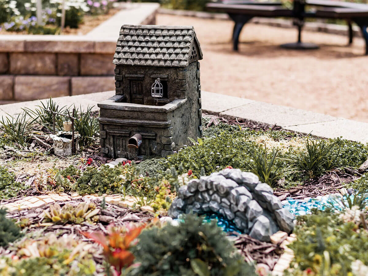 A tiny house sits in a wide, open stone planter. It's surrounded by tiny plants and vegetation. A small stone path leads to a model stone bridge, with a river of blue glass marbles underneath.